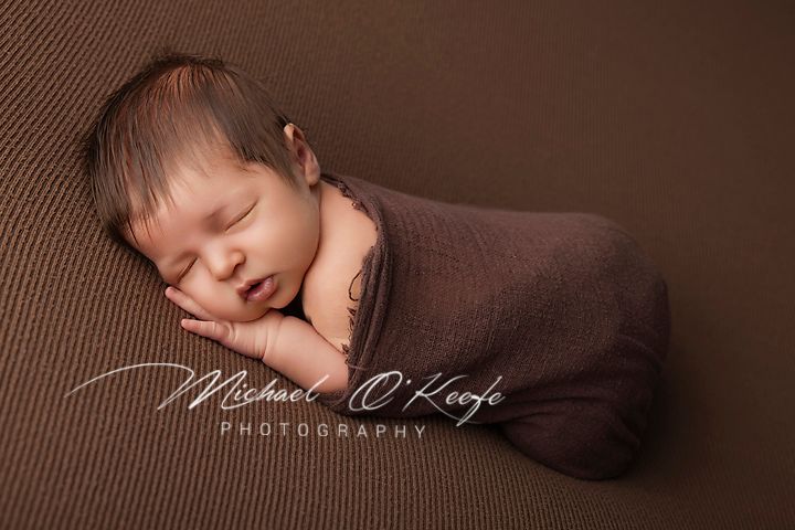 Newborn baby swaddled in brown wrap sleeping peacefully on brown textured backdrop by Michael OKeefe Photography.