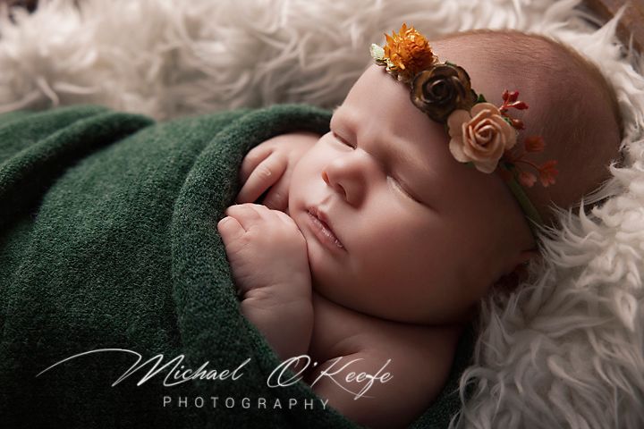 Newborn baby sleeping peacefully wrapped in green swaddle with floral headband, professional photography by Michael O'Keefe.