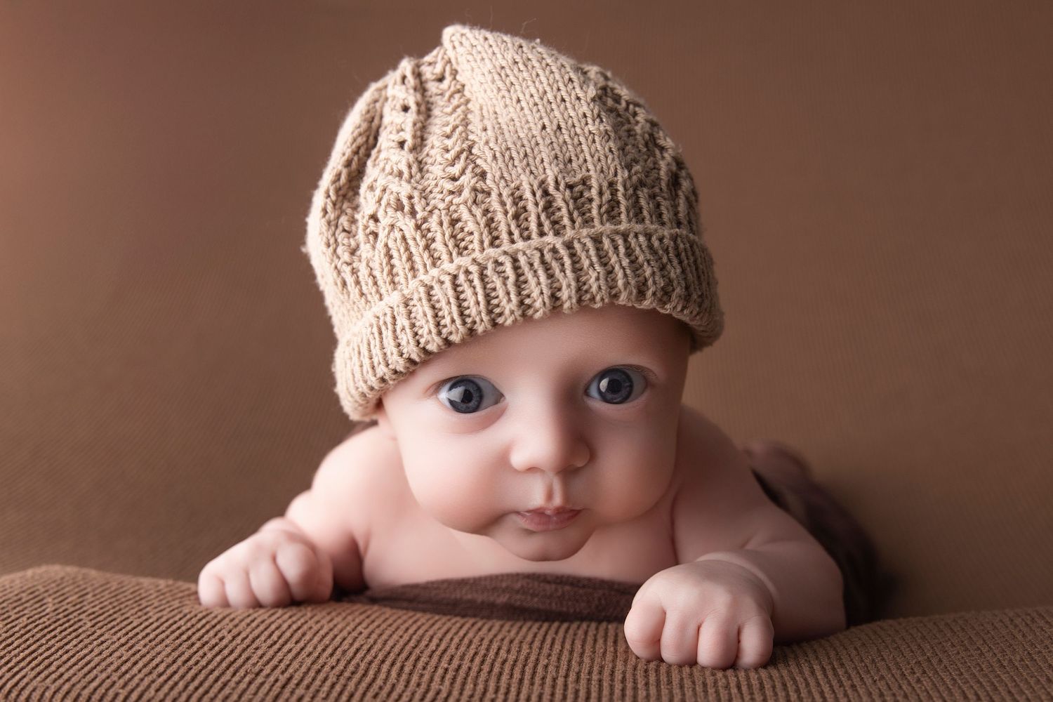 Adorable baby wearing a knit beige hat lying on a brown blanket, looking up with big blue eyes.
