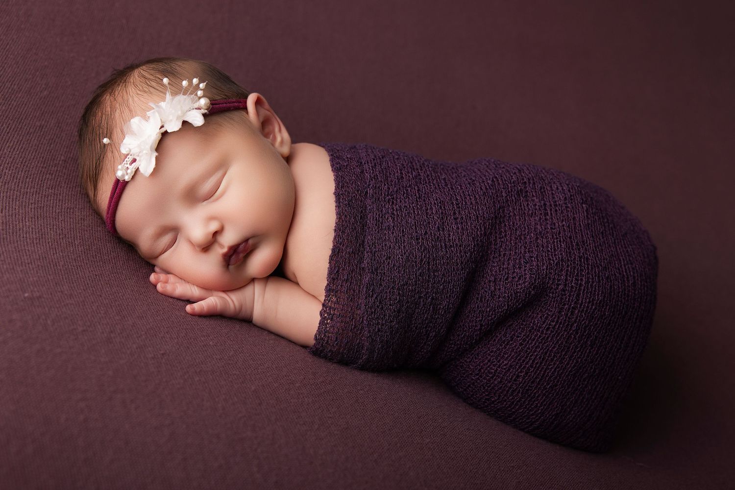 Newborn baby girl swaddled in purple wrap with floral headband, sleeping peacefully on matching purple background.
