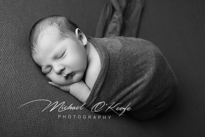Black and white newborn photo of a sleeping baby swaddled in a gray wrap by Michael O'Keefe Photography.