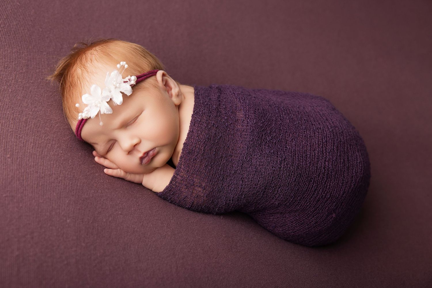 Newborn baby girl swaddled in purple wrap with floral headband, sleeping peacefully on matching purple background.