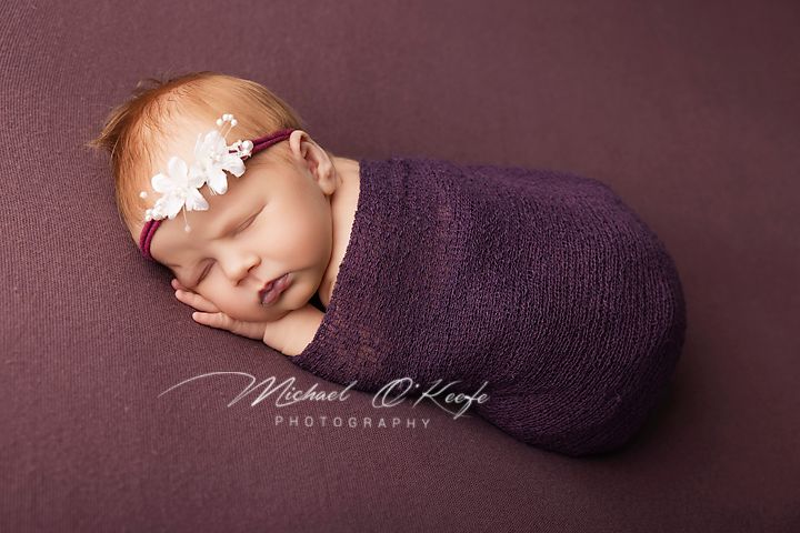 Newborn baby swaddled in purple wrap with floral headband, sleeping peacefully on purple backdrop.