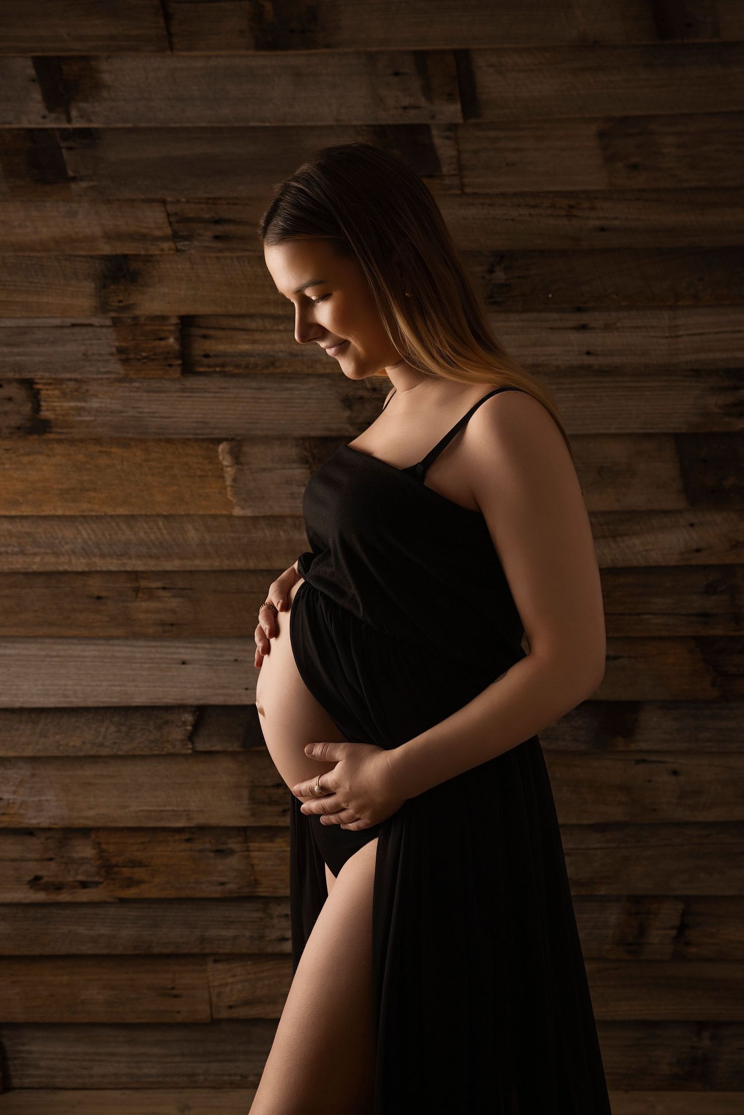 Elegant maternity silhouette in black dress posed against wooden paneled wall with moody lighting.