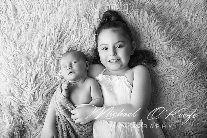 Black and white photo of a young girl smiling while holding a sleeping newborn baby on a fluffy rug.