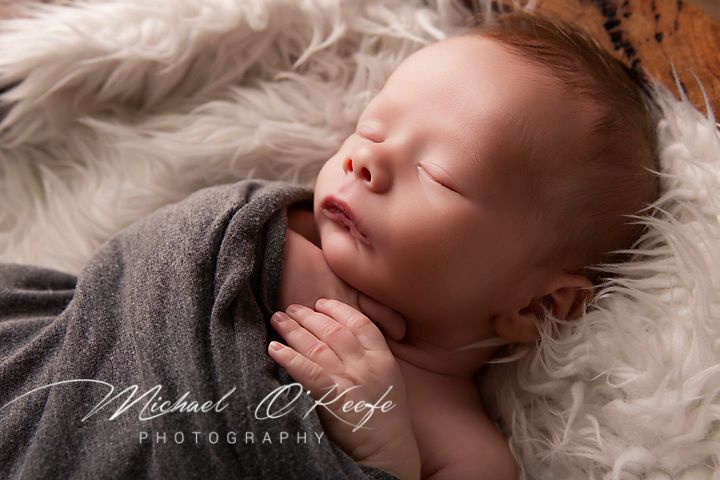 A sleeping newborn wrapped in a gray blanket rests peacefully on soft white fur.