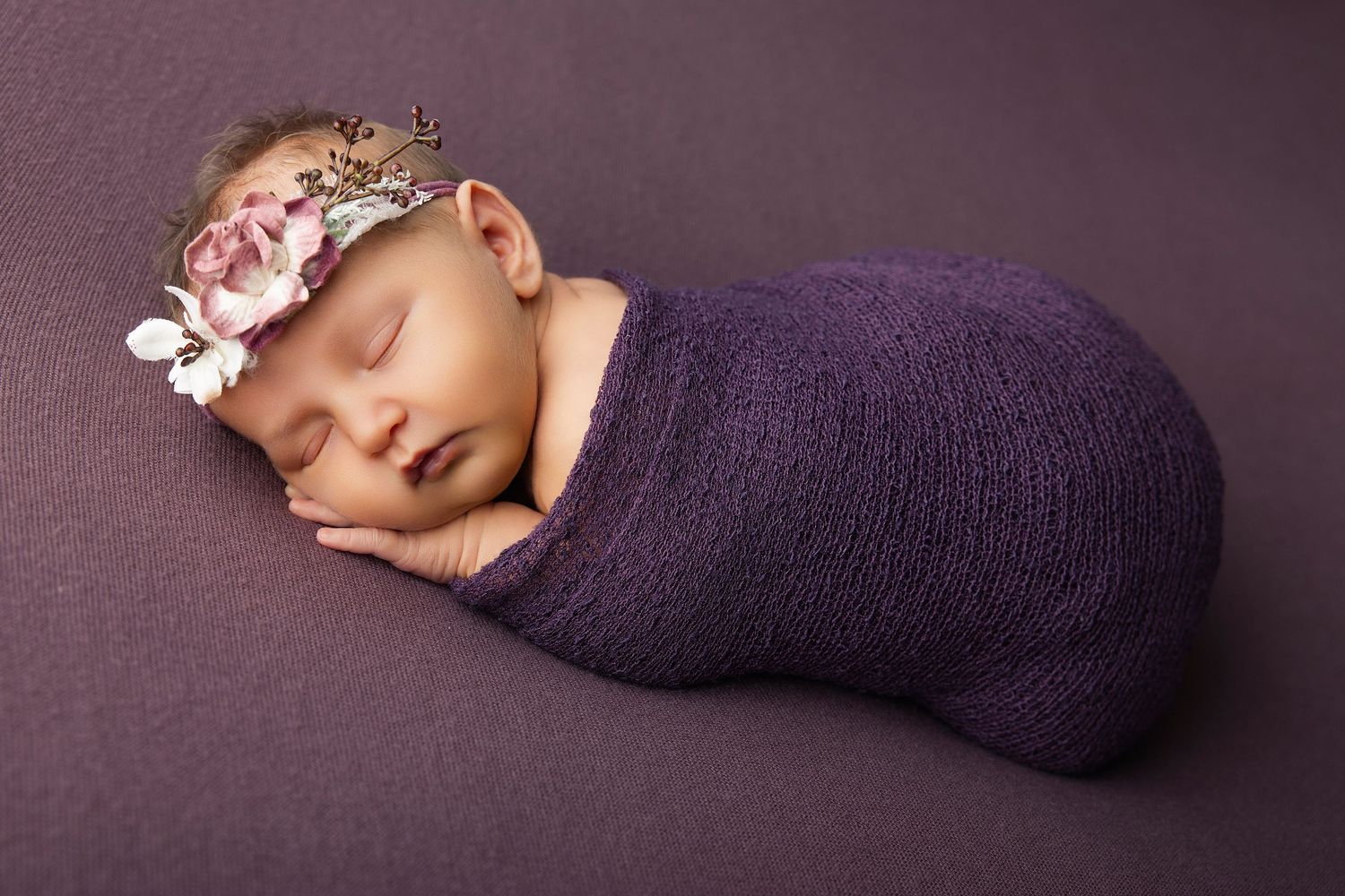 Newborn baby girl swaddled in purple wrap with floral headband, sleeping peacefully on purple background.
