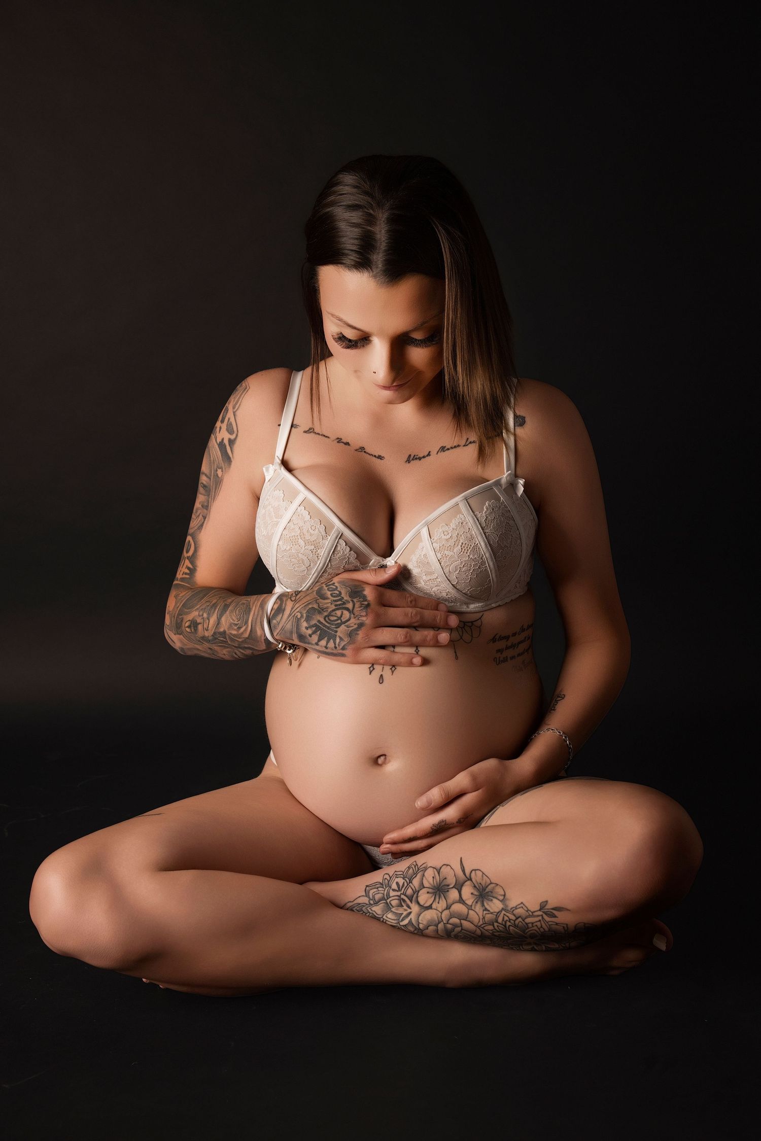 Artistic maternity portrait showing an expectant mother with tattoos in white lingerie against dark background.