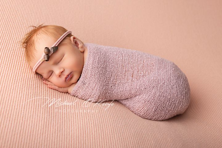 Sleeping newborn baby swaddled in pink wrap with headband on pink background.
