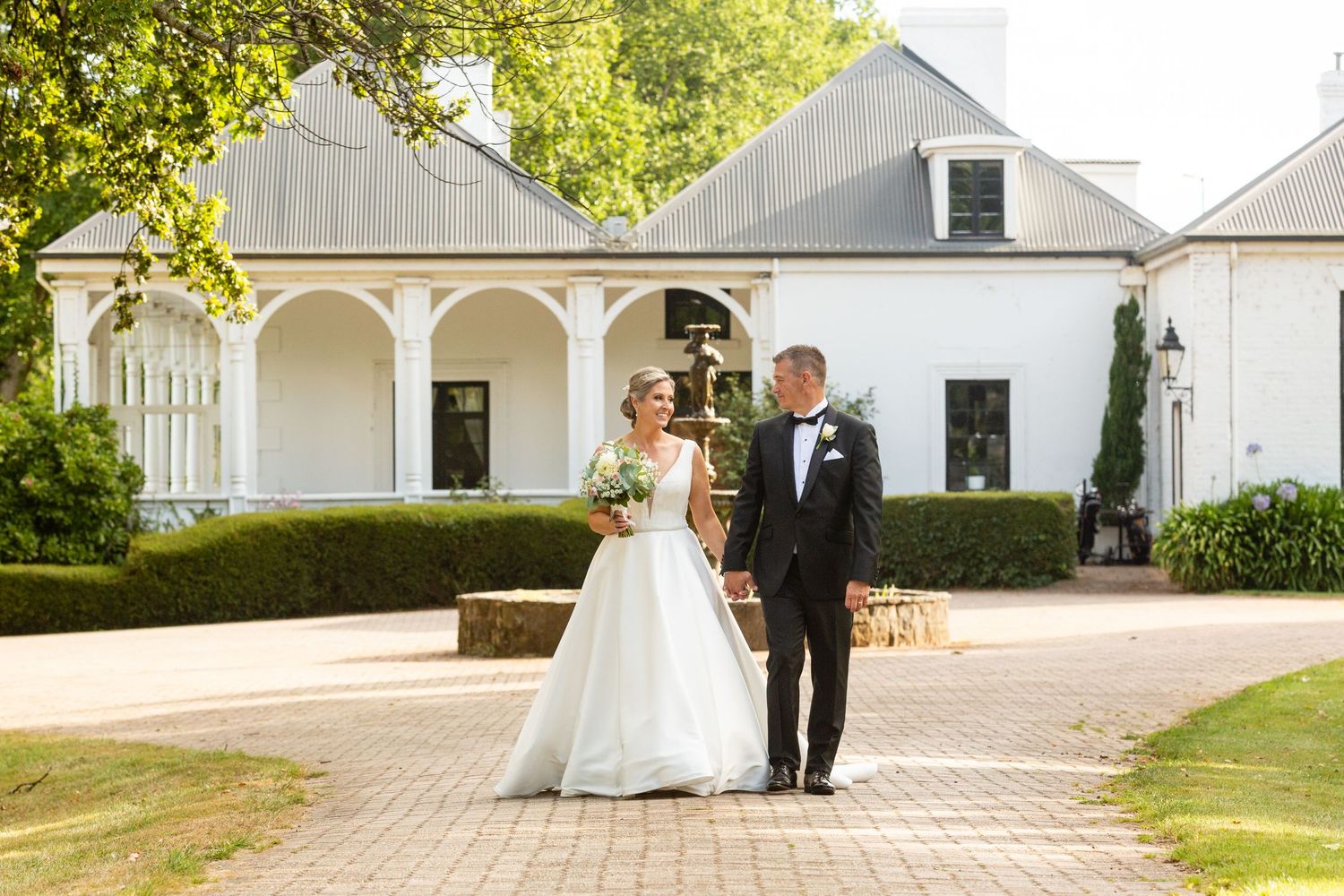 A newlywed couple walks along a brick path in front of a classic white southern-style mansion.
