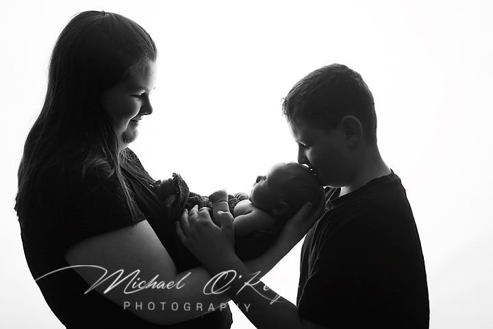 Black and white photo of a mother and older sibling lovingly holding a newborn baby by Michael OKe Photography.