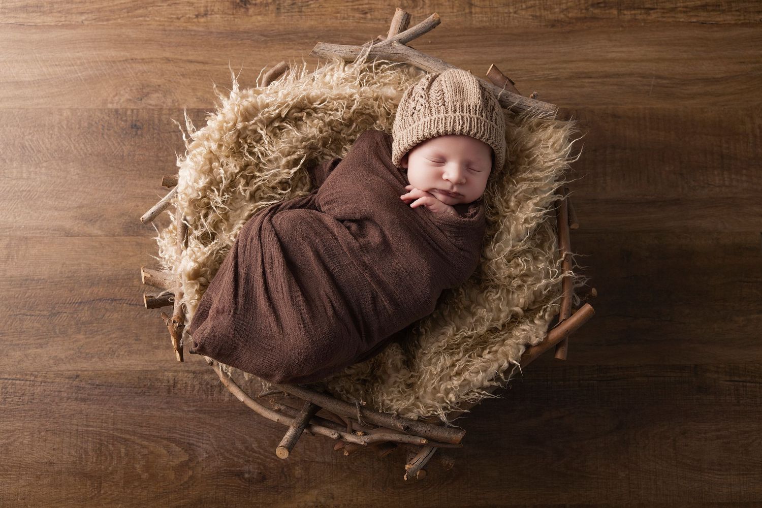 A sleeping newborn wrapped in brown swaddle lies in a cozy nest made of hay on wooden background.