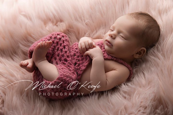 Newborn baby in pink lace outfit sleeping peacefully on soft pink fur blanket in professional studio setting.