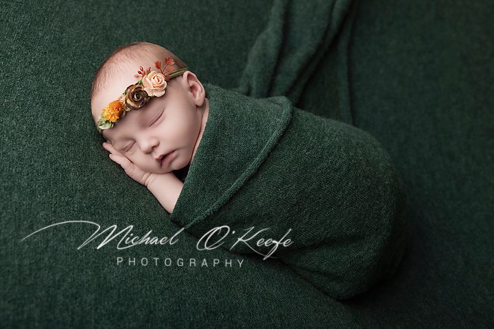 Newborn baby swaddled in green wrap with floral headband, sleeping peacefully in professional studio photo.