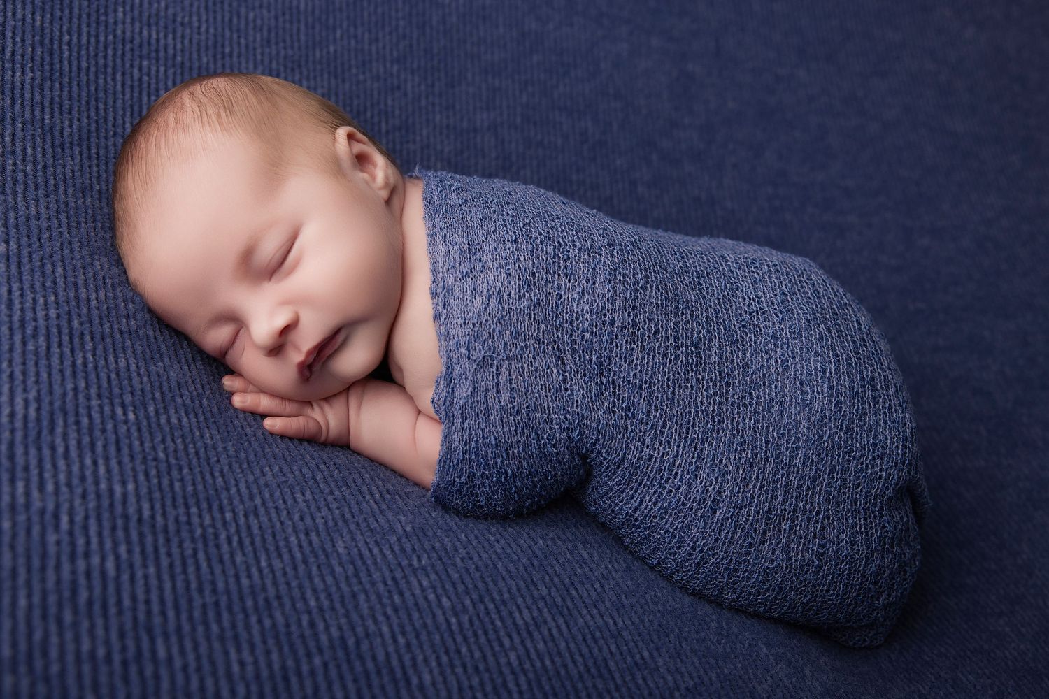 Peaceful newborn baby swaddled in a blue knit wrap, sleeping on a matching navy textured background.