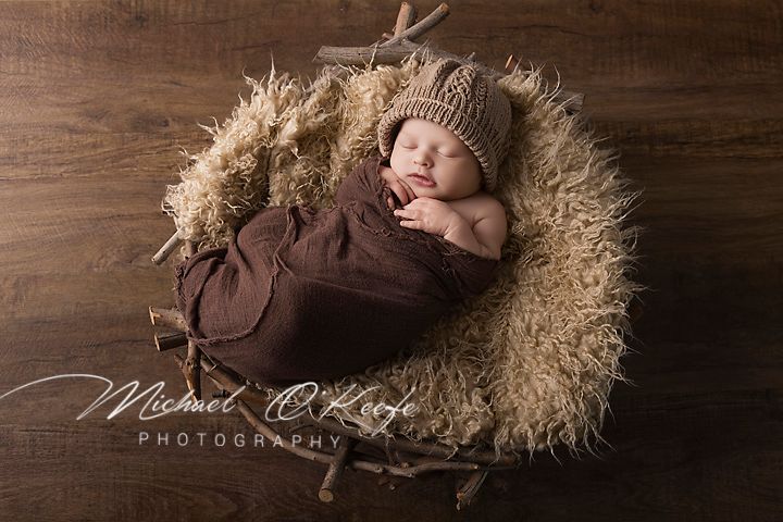 Newborn baby sleeping in a rustic wooden bowl with fluffy fur, wrapped in brown fabric, wearing a knit bear hat.
