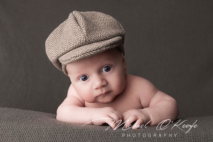 Adorable baby wearing a tan newsboy cap poses on brown cushion against dark background in studio portrait.