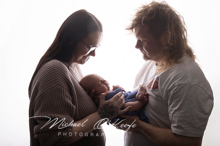 Two parents lovingly hold their newborn baby in a bright studio, captured by Michael O'Keefe Photography.