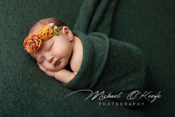 Sleeping newborn baby wrapped in green knit swaddle with orange floral headband by Michael O'Keefe Photography.