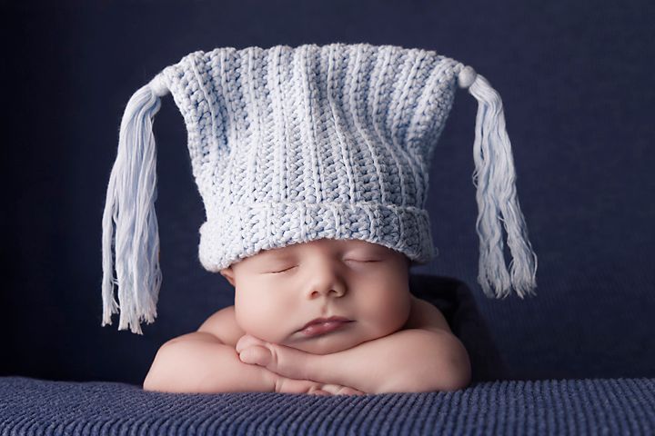 Sleeping baby wearing knit hat with tassels on blue background during newborn photoshoot.