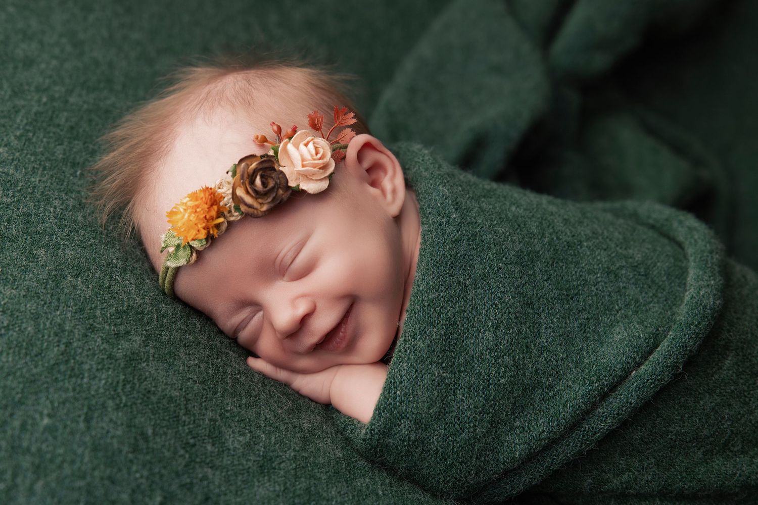 A sleeping newborn baby wrapped in dark green fabric with soft lighting creates a peaceful portrait.