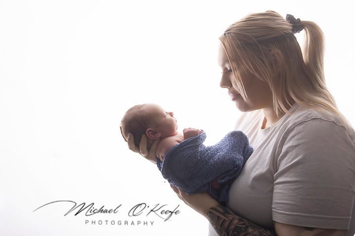 A mother tenderly holds her newborn baby wrapped in blue against a bright white background in a studio portrait.