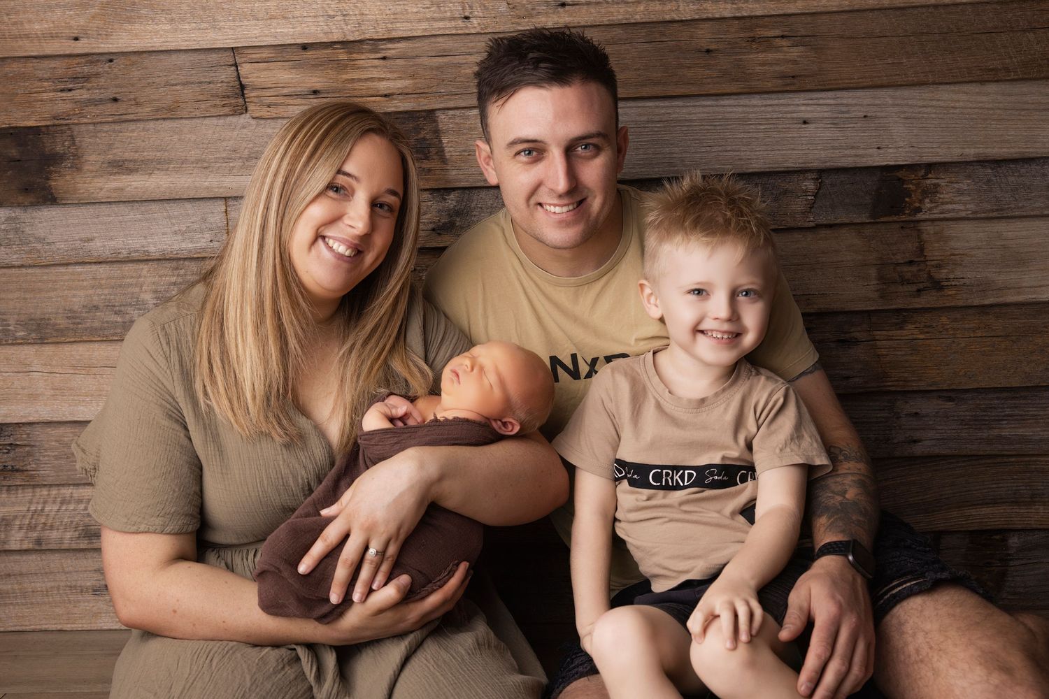Family portrait with newborn baby posed in matching brown outfits against wooden wall background.