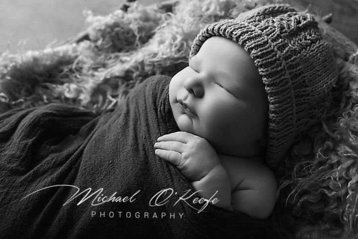 Black and white newborn photography of a sleeping baby in a knit hat, wrapped in soft fabric by Michael O'Keefe.