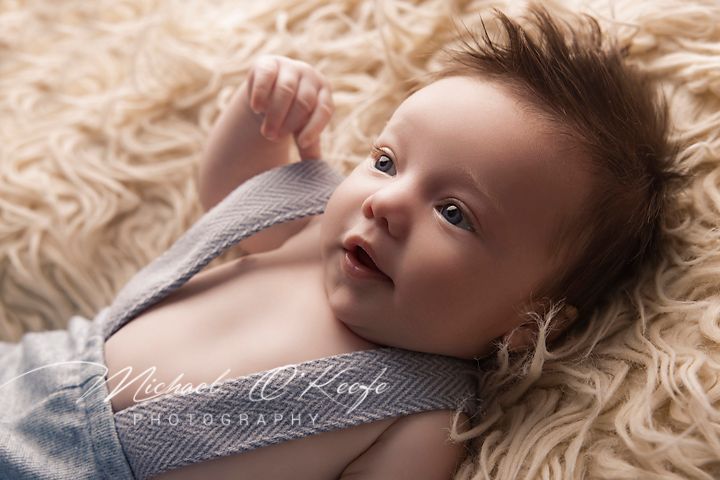 Baby in grey knit sweater lying on cream textured blanket looking up with curious expression.