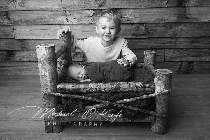 Black and white photo of a smiling toddler boy sitting with a sleeping newborn in a rustic log bed.