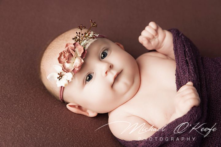 Baby wearing a delicate floral headband lies on brown backdrop during professional portrait session.