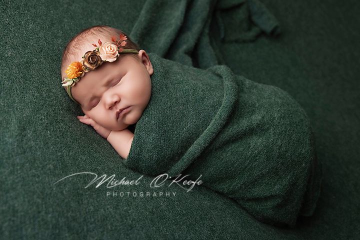 Newborn baby sleeping peacefully wrapped in dark green blanket with delicate orange flower headband.