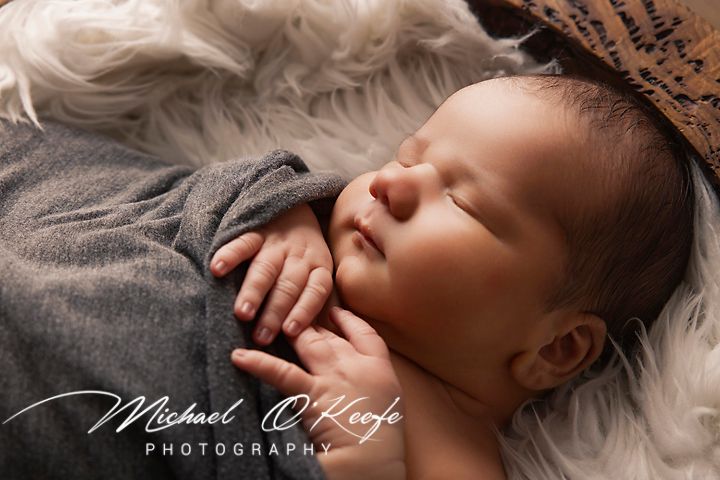 A sleeping newborn swaddled in gray fabric lies on soft white fur during a professional photography session.