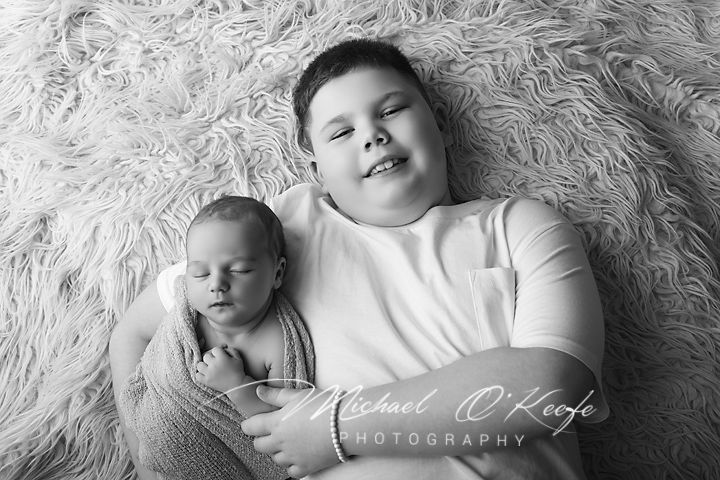 Black and white newborn photo of sleeping baby with smiling older sibling on fluffy rug by Michael O'Keefe Photography.