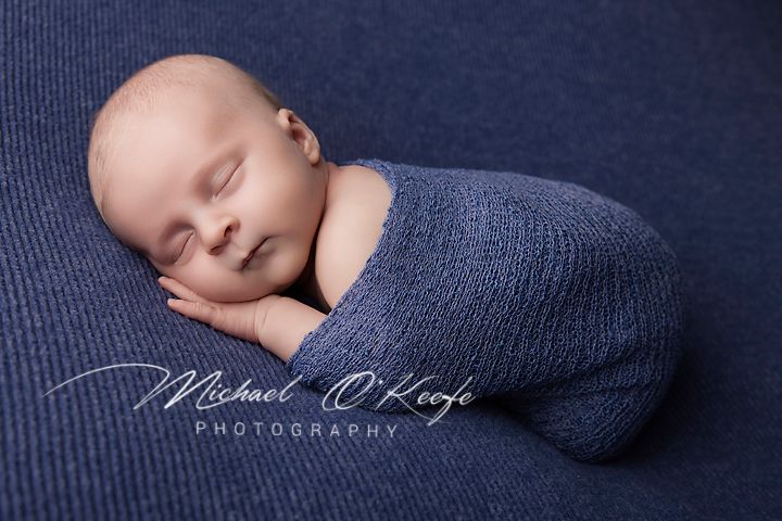 Newborn baby sleeping peacefully wrapped in a blue knit swaddle on a navy textured backdrop by Michael O'Keefe Photography.