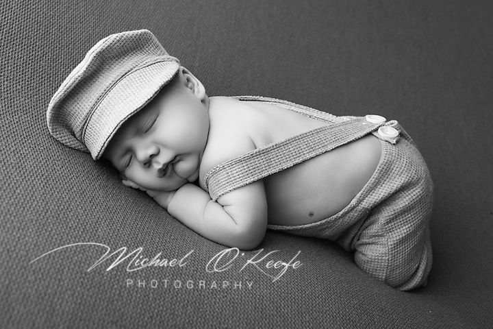 Black and white newborn photo of sleeping baby in suspenders and striped cap by Michael O'Keefe Photography.