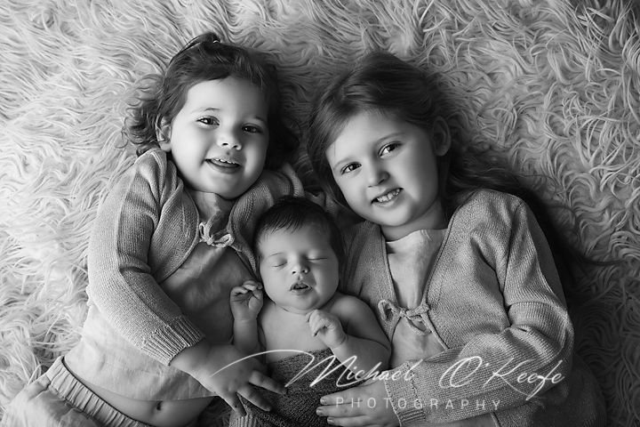Black and white photo of two young girls smiling and cuddling a sleeping newborn baby on a fluffy rug.