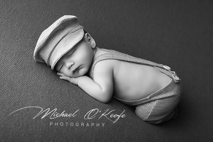 Sleeping newborn baby wearing a striped newsboy cap and suspenders, posed in black and white photography.