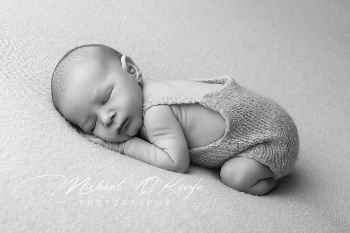 Black and white portrait of a sleeping newborn baby wearing a knit romper while lying on a textured surface.