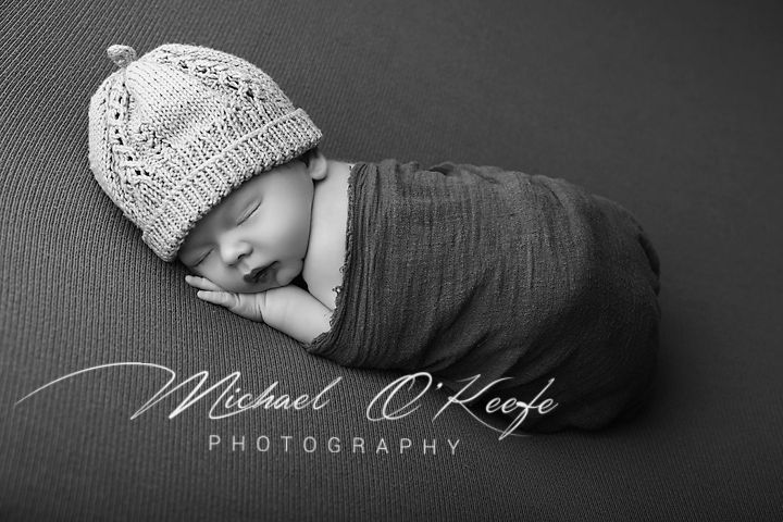 Black and white newborn photo of sleeping baby wrapped in gray swaddle wearing knit hat by Michael O'Keefe Photography.