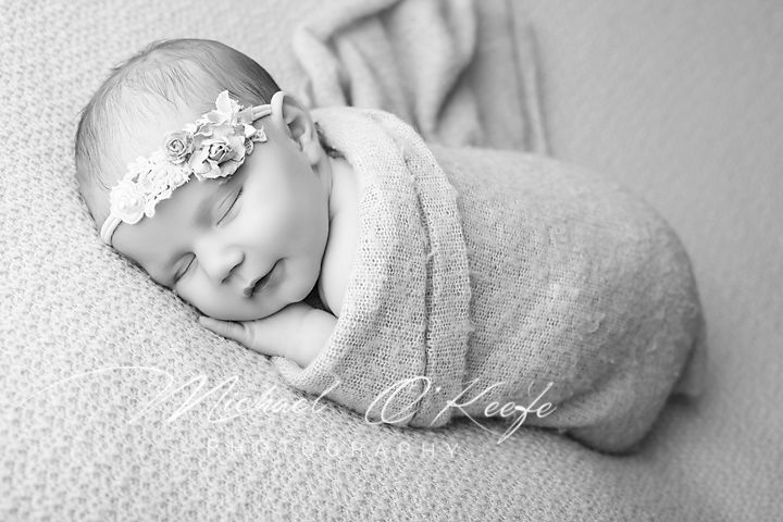 Black and white newborn photo of a sleeping baby swaddled in a soft wrap wearing a delicate floral headband.
