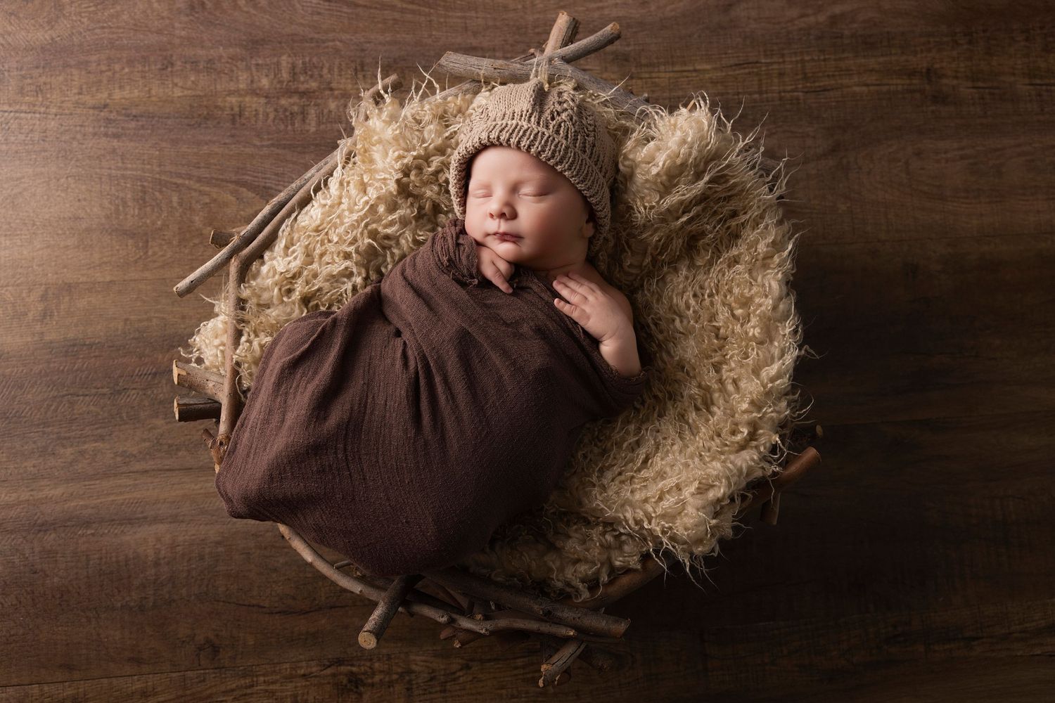 Newborn in brown wrap lying in rustic nest prop on wooden background.