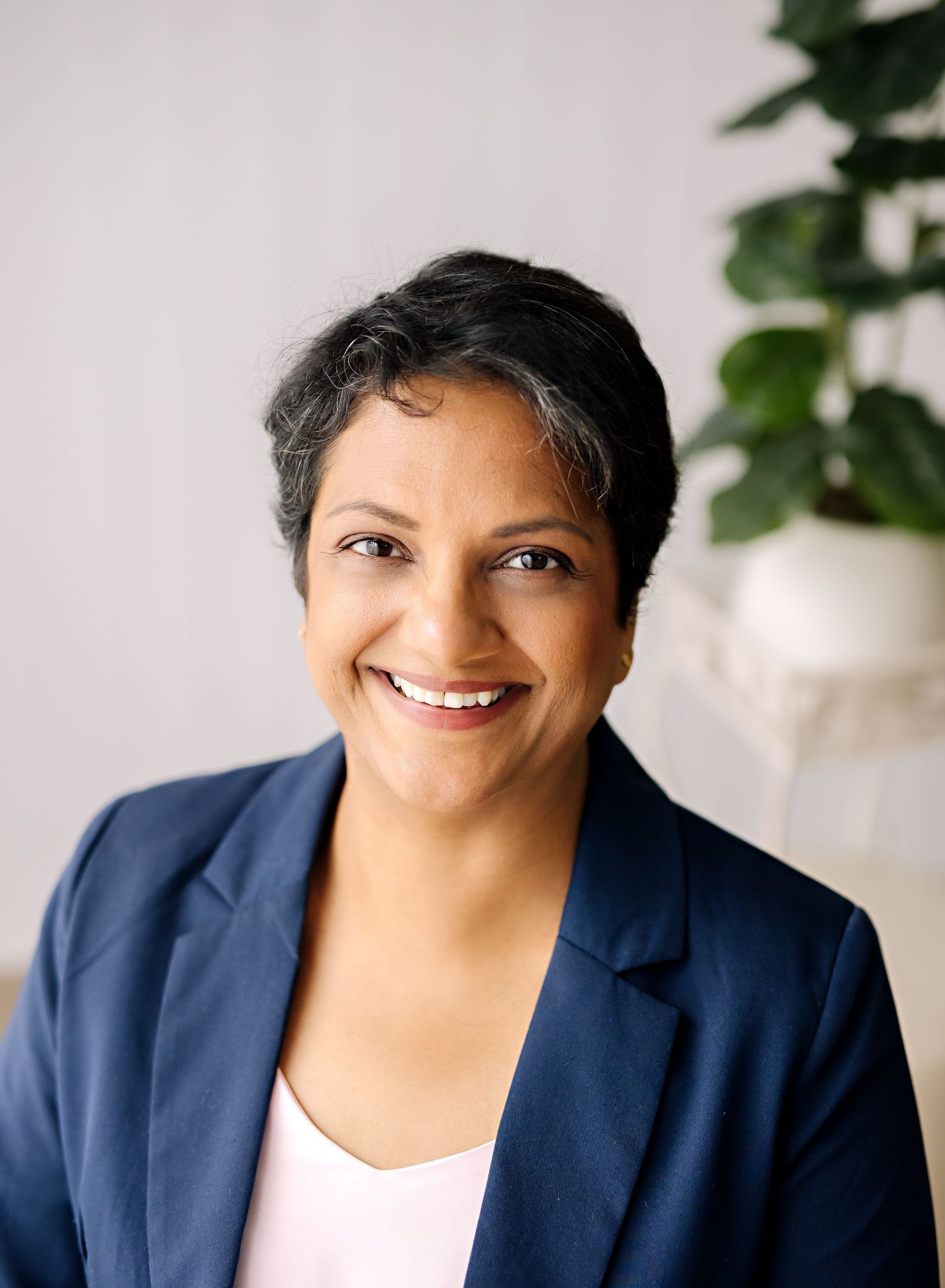 Professional wearing a navy blue blazer smiles warmly while posing against white background with green plants.