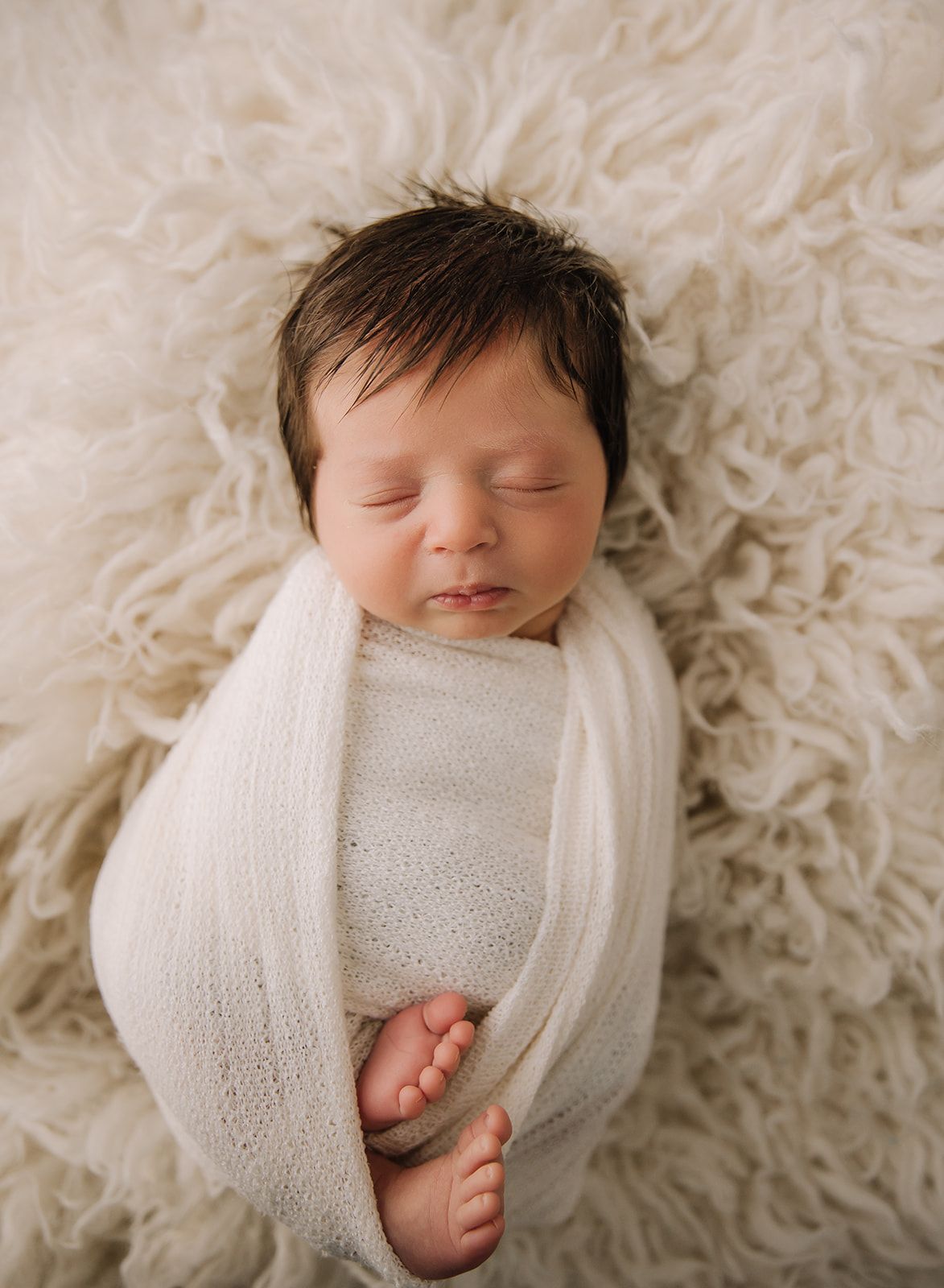 Five peaceful close-up photos of a sleeping newborn swaddled in soft white fabric against a creamy textured background.