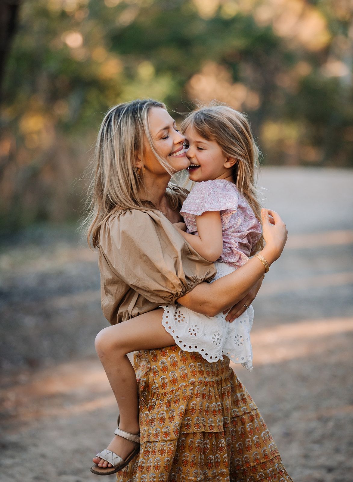 A sweet mother-daughter moment outdoors, sharing laughter and embraces in warm golden light at sunset.