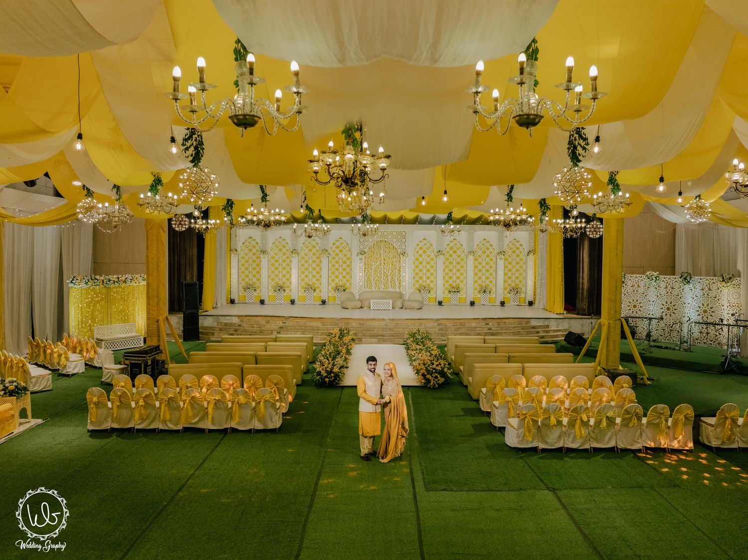 An elegantly decorated yellow wedding venue with chandeliers and rows of chairs for guests.