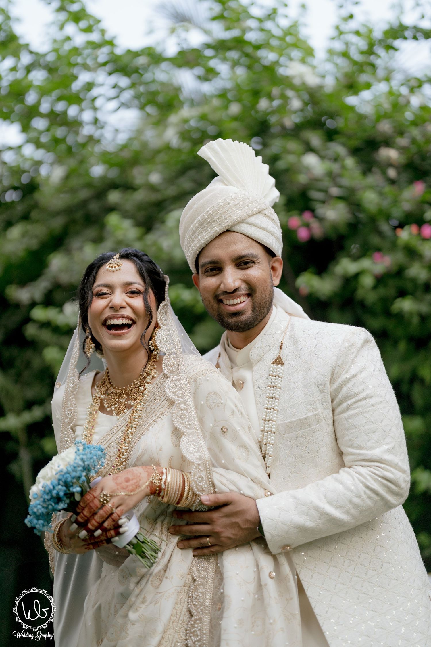 A joyful wedding couple in traditional white and cream attire share a candid moment in a garden setting.