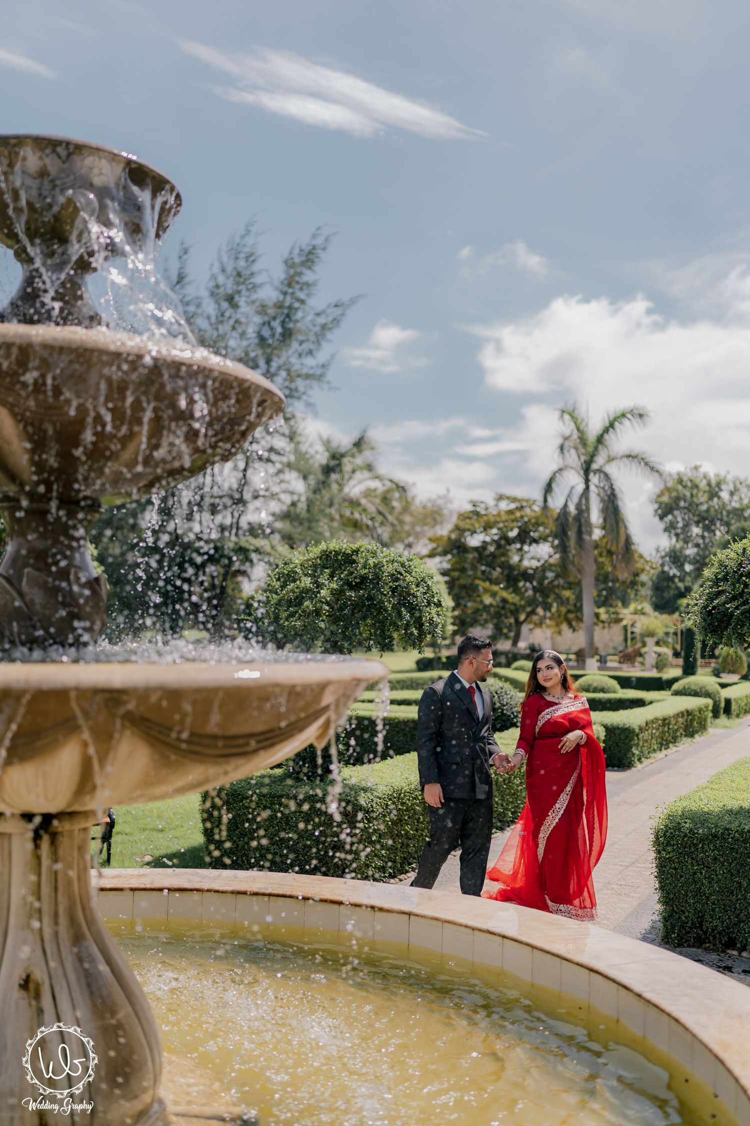 Couple walking by ornate fountain in garden, with flowing water and blue sky.