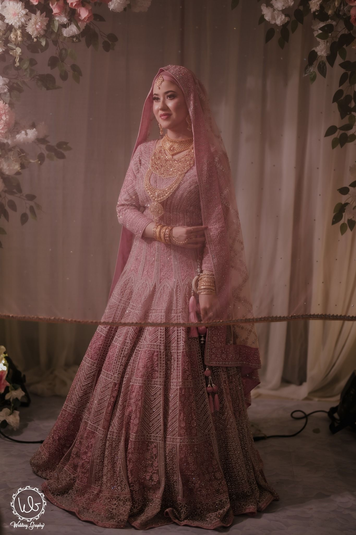 A bride in an ornate dusty pink lehenga with intricate embroidery and matching veil poses against a floral backdrop.