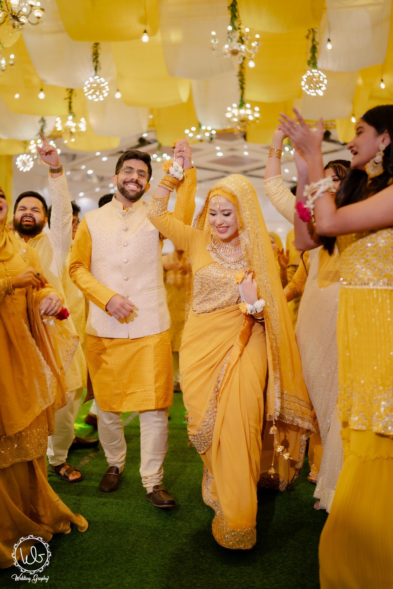 Wedding guests in yellow and gold traditional attire dance joyfully during a celebration.